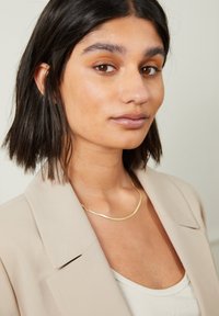 Beige blazer with wide lapels, layered over a light tank top. Gold necklace, curved and thin, contrasts against the skin tone. Short dark hair.