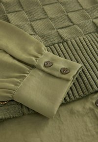 Grön texturerad tröja med ett rutmönster och ribbat avslut, med släta ärmar i silkesliknande grönt och två bruna knappar.