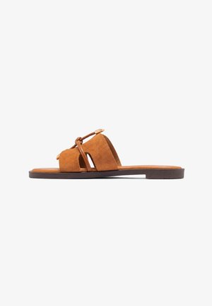 Flad slip-on sandal med brune ruskind brede stropper og tynde læder snørebånd bundet over toppen på en mørk sål.