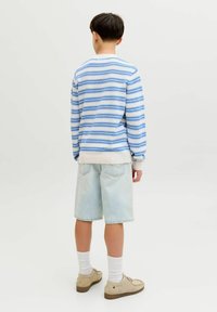 Garçon portant un pull rayé bleu et blanc, un short en jean clair, des chaussettes blanches et des chaussures beiges, debout de dos sur un fond uni.