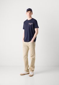 Marineblauwe T-shirt met witte logo-opdruk, beige broek en witte sneakers met bruine zolen. Pet past bij het shirt. Casual pasvorm, lichtgewicht materiaal.