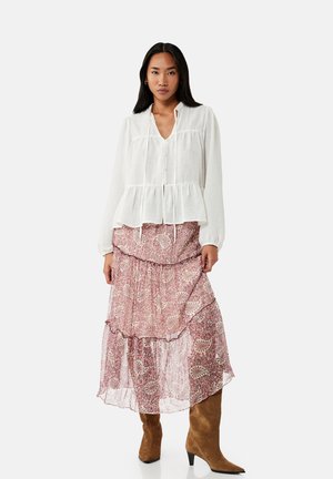 Mujer con blusa blanca de manga larga, falda midi rosa con volantes y estampado floral, y botas altas marrones de ante, de pie frente a un fondo liso.