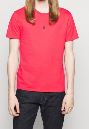 Person iført en knaldrød t-shirt med korte ærmer, med en lille mørkeblå broderet logo midt på brystet og mørkeblå jeans.