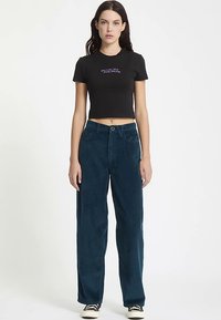 T-shirt noir coupé avec texte violet, associé à un pantalon large en velours côtelé bleu sarcelle, doté d'une fermeture à boutons et d'une texture côtelée verticale.