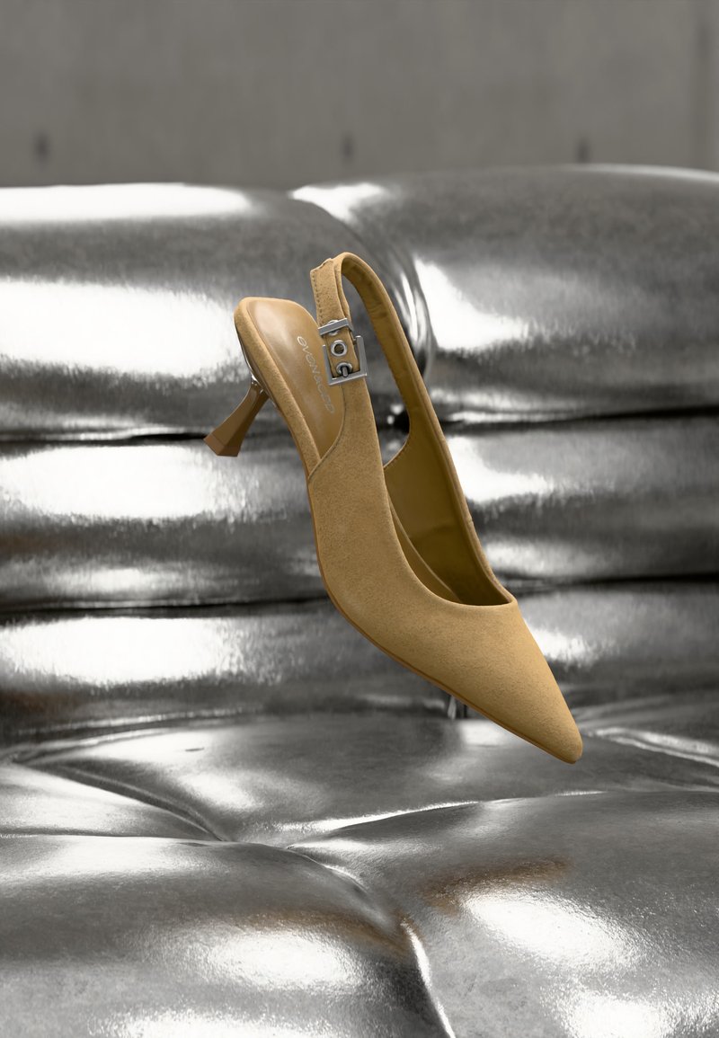 Chaussure beige en daim à talonkitten et bride arrière avec une boucle argentée, positionnée au-dessus d'une surface capitonnée métallique brillante.