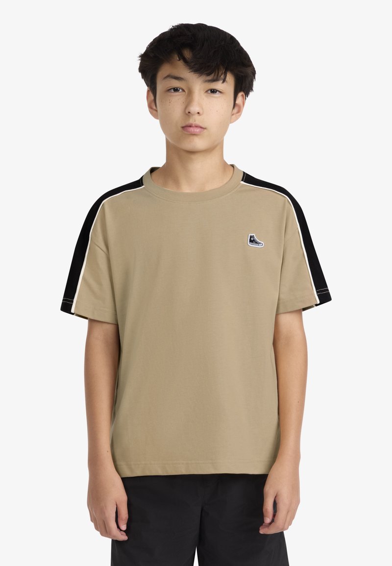 Μπεζ κοντομάνικο μπλουζάκι με μαύρα μανίκια και λευκές ρίγες. Διαθέτει μικρό λογότυπο παπουτσιού στο στήθος· βαμβακερό ύφασμα, χαλαρή εφαρμογή.