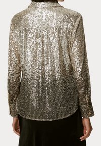 Achteraanzicht van een persoon in een lange mouwen, gouden paillettenjacket met knoopmanchetten en een zwarte rok.