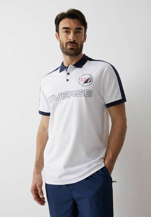 Mężczyzna z brodą w białej i granatowej koszulce polo z napisem "Diverse", stojący z jedną ręką w kieszeni na jednolitym tle.