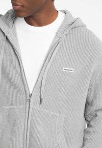 Sudadera de punto gris con cremallera y una etiqueta blanca en contraste en el lado izquierdo del pecho, con un patrón texturizado y capucha con cordón.