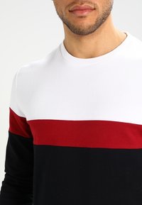 Camisa de manga larga con un diseño color block que incluye bandas en blanco, rojo y negro. La tela es suave y tiene un cuello redondo.