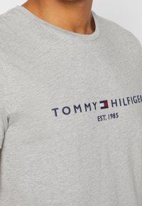 Camiseta de algodón gris con cuello redondo, con texto bordado en azul marino "TOMMY HILFIGER" y logo. Textura simple y suave.