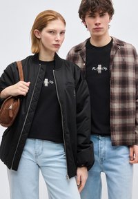 Μια γυναίκα και ένας άντρας στέκονται δίπλα-δίπλα φορώντας καθημερινά ρούχα: μαύρα μπλουζάκια με σχέδια, ανοιχτό μπλε τζιν, μαύρο μπουφάν, καφέ καρό πουκάμισο και καφέ τσάντα.