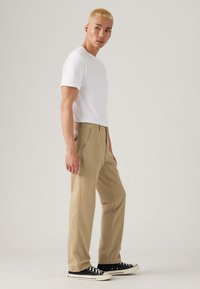 Beige broek met rechte pijpen, gecombineerd met een wit shirt met korte mouwen. Zwarte sneakers met witte zolen, staande tegen een lichte achtergrond.