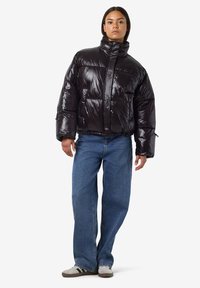 Juoda pūkinė striukė su aukštu apykakle, blizgia tekstūra ir horizontaliu siuvinėjimo raštu; dėvima su laisvais mėlynais džinsais ir laisvalaikio sportbačiais.