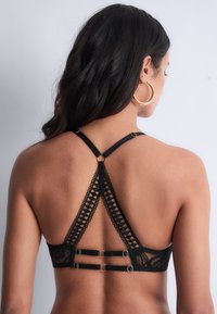 Femme montrant le dos en portant un bralette en dentelle noire avec un design géométrique et de grandes boucles d'oreilles en or, sur fond uni.