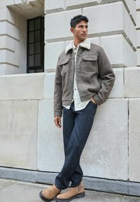 Braune Wollmischung Jacke mit einem cremefarbenen Pelzkragen, zwei Brusttaschen, getragen über einem karierten Hemd, kombiniert mit dunklen Jeans und hellbraunen Stiefeln.