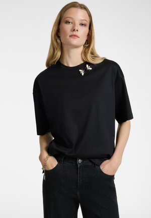 T-shirt nera oversize a maniche corte, con due spille decorative a forma di ape sul colletto, abbinata a jeans scuri.