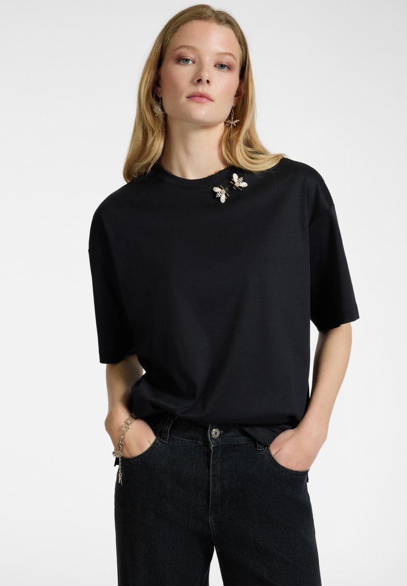 T-shirt nera oversize a maniche corte, con due spille decorative a forma di ape sul colletto, abbinata a jeans scuri.
