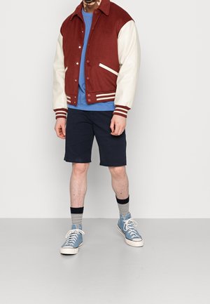 Červeno-béžová univerzitní bunda s límečkem, doplněná modrým tričkem, tmavě modrými šortkami a modrými teniskami s bílými tkaničkami a pruhovanými ponožkami.