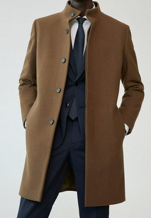 Homme portant un manteau en laine marron boutonné par-dessus un costume bleu marine avec une chemise blanche et une cravate bleu marine à motifs, les mains dans les poches du manteau.