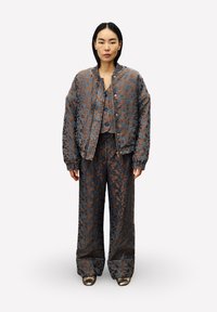 Vrouw in een gecoördineerde oversized jas, vest en wijde broek met een bruin en blauw bloemenpatroon, met metallic schoenen, staand tegen een witte achtergrond.