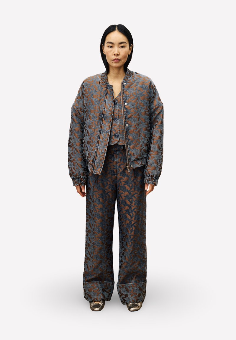 Vrouw in een gecoördineerde oversized jas, vest en wijde broek met een bruin en blauw bloemenpatroon, met metallic schoenen, staand tegen een witte achtergrond.