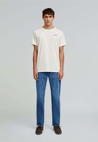 Off-white bomulds T-shirt med korte ærmer, parret med blå lige ben jeans og brune snørebåndssko. Simpelt design, uden synlige mønstre.