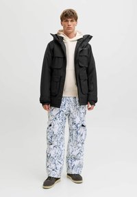 Sort vandtæt jakke med flere frontlommer over en lys beige hættejakke; parret med hvide bukser med blå marmorering.