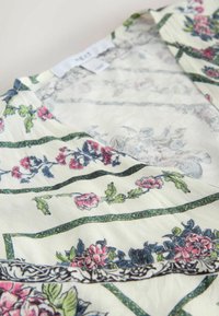 Balts audums ar rozā un zilām ziedu rakstiem un zaļām vīnogulāju lapām, tuvplāns uz apģērba kakla izgriezumu un etiķeti.