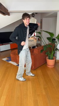 Mladi muškarac se smiješi i oblači crnu jaknu u modernom dnevnom boravku s drvenim podom, narančastim sofom i velikom sobnom biljkom u posudi.