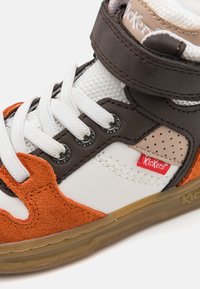 Sapatilha alta com uma combinação de couro branco, camurça castanha e detalhes laranja. Inclui painéis em malha, atacadores e faixa de Velcro.