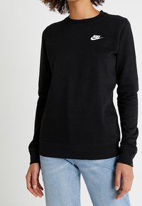 Svart sweatshirt i mjukt tyg, med rund hals, långa ärmar och en vit Nike-logotyp på övre vänstra bröstet.