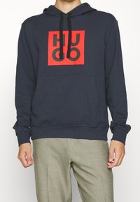 Persona vestida con una sudadera navy con cordones negros y un logo cuadrado rojo con letras abstractas negras, combinada con pantalones gris claro.