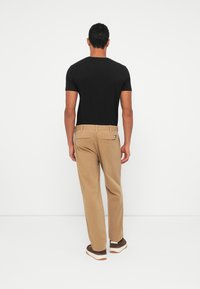 Muž oblečený v čiernom tričku s krátkym rukávom, béžových nohaviciach so bočnými vreckami a hnedých teniskách stojí voči bielej stene, pohľad zozadu.