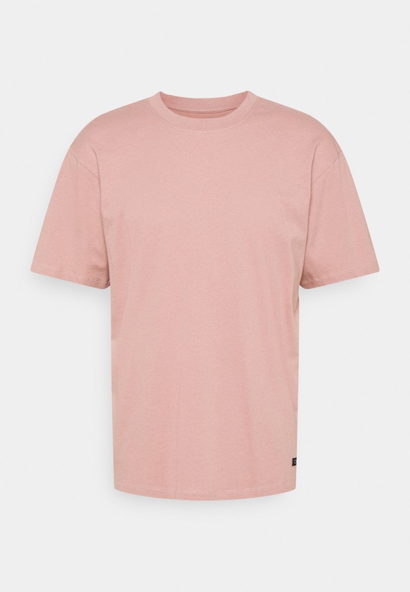 T-shirt simple rose clair à manches courtes et col rond avec une petite étiquette noire près de l'ourlet inférieur sur un fond blanc uni.
