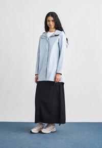 Svetlomodrá nepremokavá bunda so zipsom vpredu, kapucňou a dlhými rukávmi. Kombinovaná s čiernou dlhšou sukňou a sivými športovými topánkami.