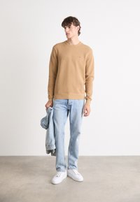 Maglione marrone chiaro con scollatura a girocollo, jeans azzurri e sneakers bianche. Il modello tiene in una mano una giacca di denim chiaro.
