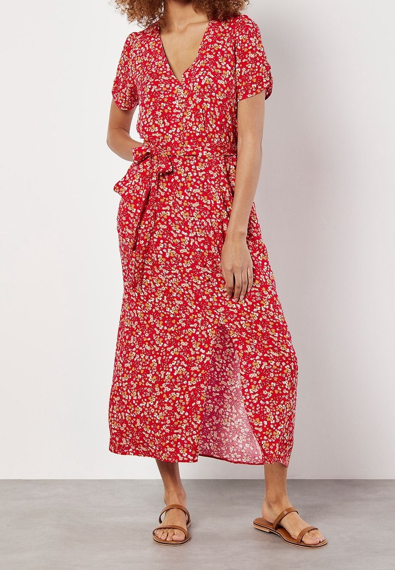 Robe maxi fleurie rouge avec manches courtes, détail de ceinture à nouer, et une silhouette fluide. Présente de petits motifs de fleurs blanches et jaunes.