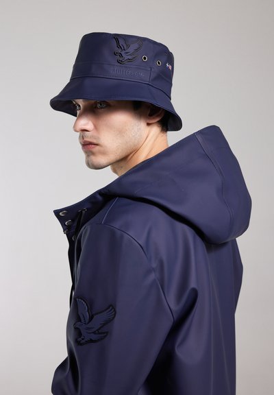 Marineblaue Bucket-Haube und Regenjacke aus glattem, wasserdichtem Material. Die Jacke hat ein Adler-Logo auf dem Ärmel und Druckknopfverschlüsse.