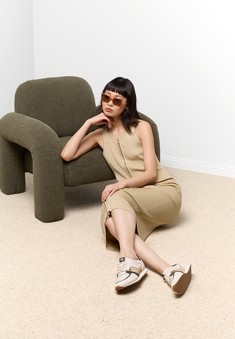Robe sans manches en tricot beige avec une fermeture éclair ; chaussures de sport marron et crème ; lunettes de soleil oversize d'un beige clair. Chaise texturée en vert olive.