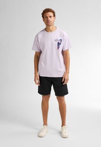 Lichtpaarse t-shirt met een grafisch ontwerp op de borst, gecombineerd met zwarte shorts en witte sneakers. Katoenen stof, relaxte pasvorm.
