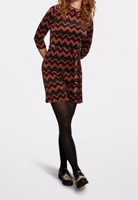 Kortärmad klänning med zigzag-mönster i röd, grön och svart, knuten i midjan. Bärs med svarta strumpbyxor och tvåfärgade skor.