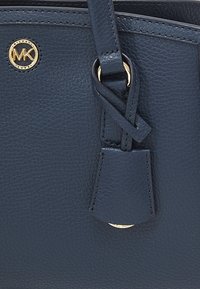 Détail d'un sac à main en cuir texturé bleu marine avec le logo Michael Kors doré et une sangle en cuir nouée attachée à un anneau en or.