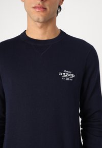 Sweat-shirt bleu marine en tissu texturé, comprenant un col rond et le logo Tommy Hilfiger brodé en blanc sur la poitrine gauche.