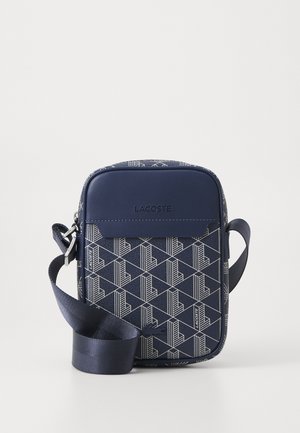 Sac bandoulière bleu marine avec motif géométrique blanc, poche avant, logo Lacoste embossé et bandoulière réglable.