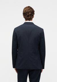 Σκούρο μπλε σακάκι και παντελόνι, σε εφαρμοστή γραμμή, με μονό άνοιγμα στο πίσω μέρος, λείο ύφασμα και λεπτομέρεια με τέσσερα κουμπιά στο μανίκι. Χωρίς ορατή επωνυμία.