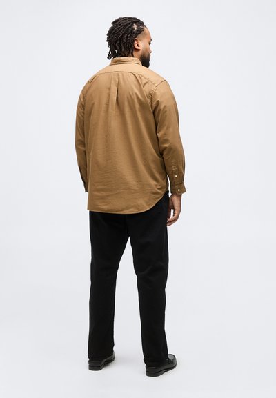 Άνδρας με ντρέντλοκς στέκεται πλάτη, φοράει καφέ μακρυμάνικο πουκάμισο, μαύρο παντελόνι και μαύρα παπούτσια σε απλό φόντο.