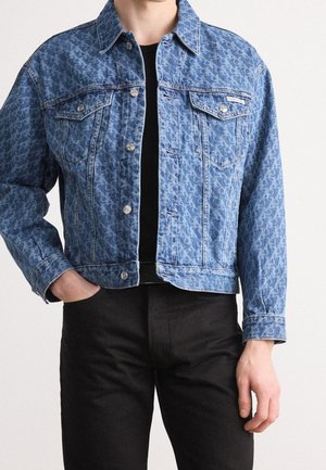 Osoba ubrana w niebieską, wzorzystą kurtkę dżinsową z srebrnymi guzikami, na czarnej koszuli i czarnych spodniach, z jedną ręką w kieszeni.