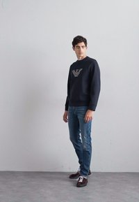 Sudadera negra con logo texturizado en el pecho, combinada con jeans de color azul claro. El modelo lleva zapatillas marrones, de pie sobre un fondo neutro.