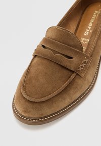 Mocassin en daim marron avec bride à monnaie, coutures détaillées et bout arrondi sur un fond clair.